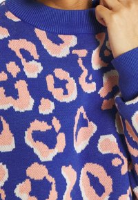 Pull en tricot bleu avec des taches léopard roses et blanches, col et poignets côtelés, porté par une personne touchant son cou avec une main.