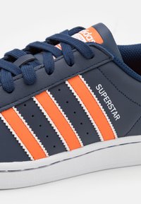 Modré kožené tenisky se třemi oranžovými pruhy, bílými zigzagovými detaily, perforacemi pro prodyšnost a vyšívaným logem "SUPERSTAR".