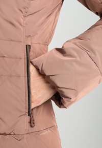 Chaqueta acolchada en un tono rosa apagado con un bolsillo lateral con cremallera, detalle de puño acanalado y textura de tejido suave y ligero.