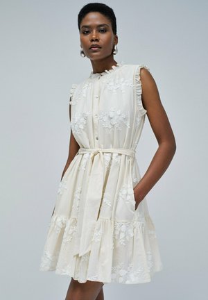 Robe en coton beige clair avec broderies florales, manches volantées, ouverture à boutons sur le devant, ceinture à la taille et jupe évasée, avec poches latérales.