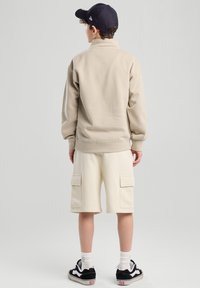 Beige fleece pullover with a high collar, paired with cream cargo shorts featuring front pockets, and black sneakers with white accents.