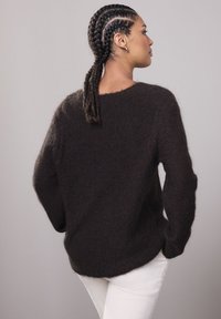 Naine punutud juustega ja kuldsete rõngaskõrvarõngastega, seljas tume karvane kampsun ja valged püksid, seisab külje peal käed taskutes.