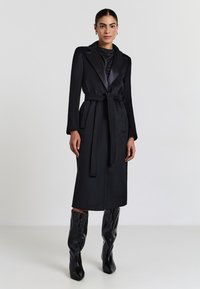 Cappotto in lana nera con cintura, maniche lunghe e rever in satin. Abbinato a stivali neri al ginocchio lucidi, con una texture liscia.