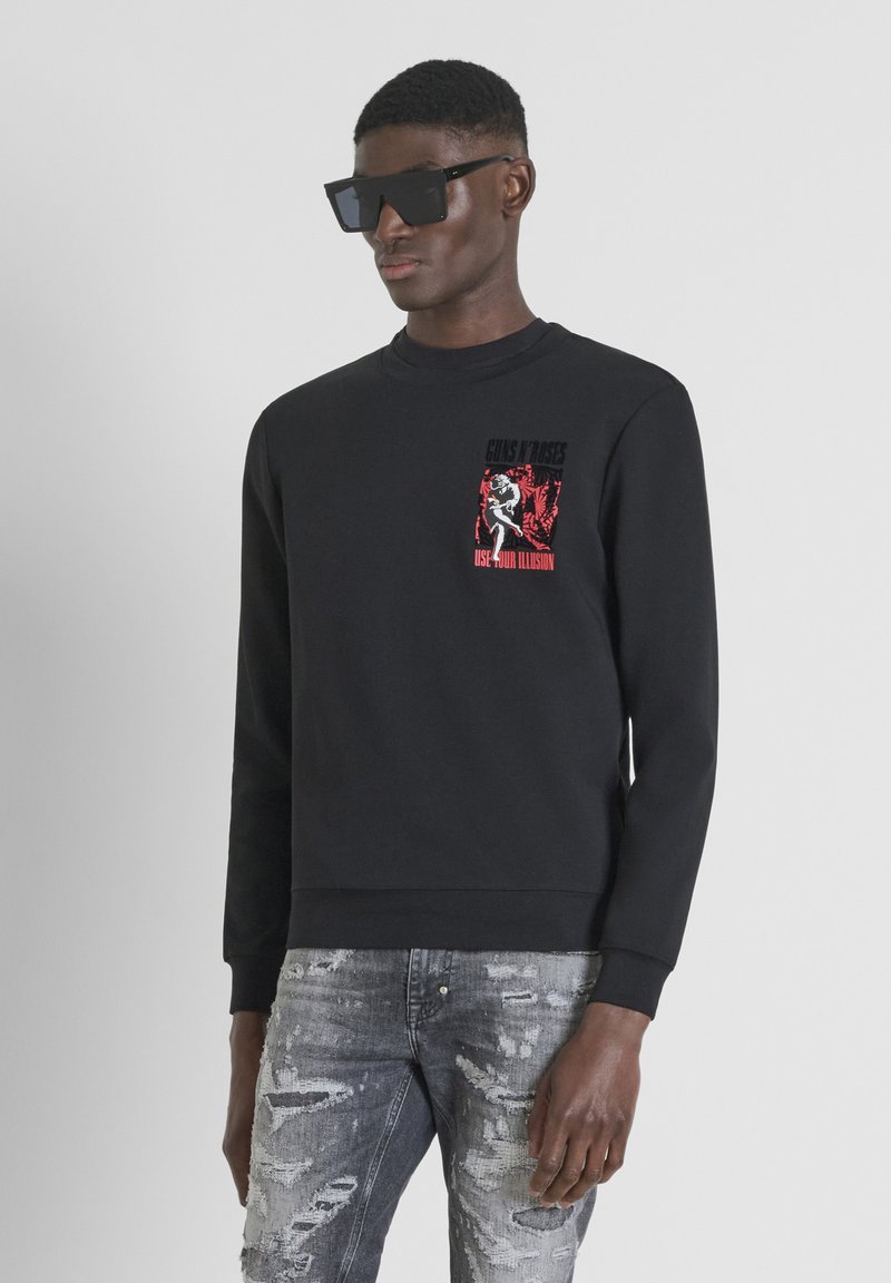 Sudadera negra de algodón con cuello redondo. Presenta una impresión gráfica en rojo y blanco en el área del pecho izquierdo. Combinada con jeans grises desgastados.