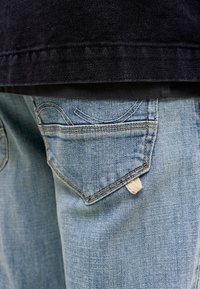 Οπίσθια όψη τσέπης ανοιχτό μπλε τζιν με καμπύλες ραφές και μια μικρή λευκή υφασμάτινη ετικέτα, φορεμένη με σκούρο τζιν μπουφάν.