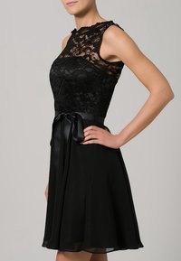 Femme portant une robe noire en dentelle sans manches avec un corsage ajusté, un décolleté transparent, un ruban de satin à la taille et une jupe fluide longueur genou, main sur la hanche.