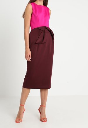 Vrouw die een mouwloze roze top draagt met een donker bordeauxrode kokerrok met hoge taille en oranje sandalen met hoge hakken, staand op een witte vloer.