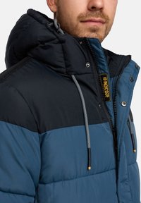 Wattierte Jacke mit Kapuze, zweifarbiger Gestaltung in Dunkelblau und Schwarz, mit grauen Kordeln und gelber Markenkennzeichnung am Reißverschluss.