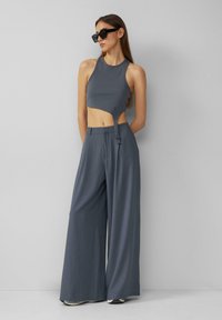 Šedý žebrovaný crop top s bočním vykrojením v kombinaci s volnými šedými širokými kalhotami. Model má na sobě černé sluneční brýle. Hladká, strukturovaná tkanina.