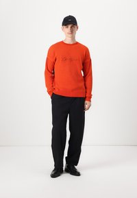 Maglione arancione a coste con testo ricamato, abbinato a pantaloni neri e scarpe nere. Il modello indossa un cappello nero, in piedi su uno sfondo bianco.