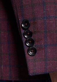 Tkanina w kratę w kolorze bordowym i niebieskim z teksturowanym splotem. Cztery czarne guziki na mankiecie, każdy z małym otworkiem w centrum.