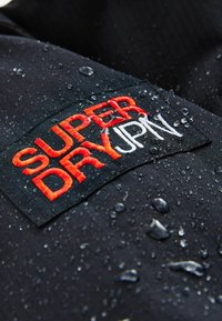 Tessuto nero con una finitura testurizzata, caratterizzato da un'etichetta ricamata "SUPER DRY" in rosso e bianco, con gocce d'acqua distribuite sulla superficie.