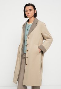 Manteau beige avec un col texturé à motif foncé. Il présente des manches longues, des boutons à l'avant et des poches latérales. Porté par-dessus une chemise bleue boutonnée.