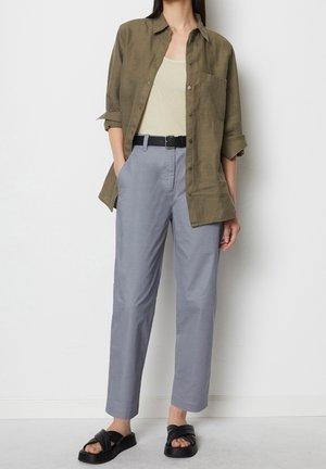 Femme portant une chemise boutonnée olive sur un débardeur crème, un pantalon taille haute gris clair avec une ceinture noire et des sandales à plateforme noires.