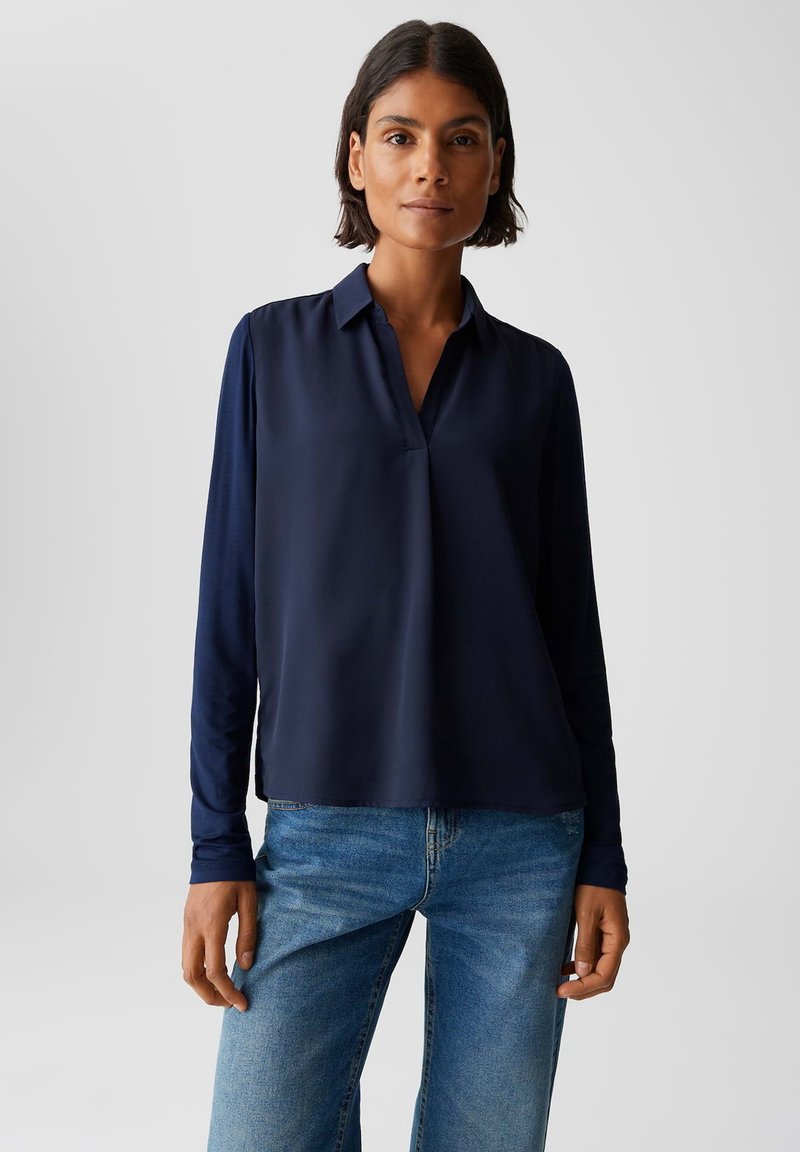 Marineblaue Bluse mit langen Ärmeln, klassischem Kragen und lockerer Passform; glatte Textur und gerader Saum. Kombiniert mit hellblauen Jeans.