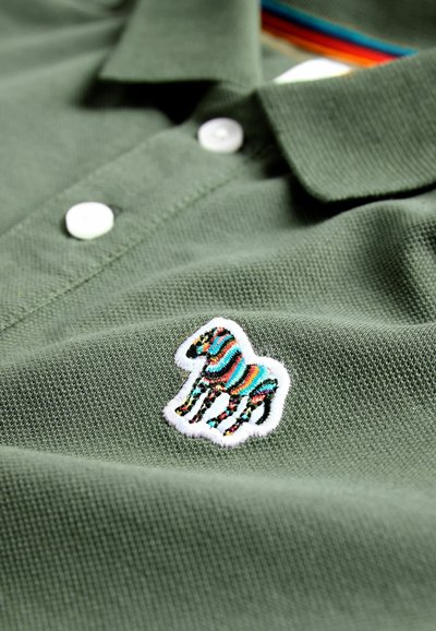 Khaki polo marškiniai su balta siuvinėta zebra uždanga, papuošta spalvingomis juostelėmis. Ant apykaklės matomi du balti mygtukai. Tekstūruota medvilnė.