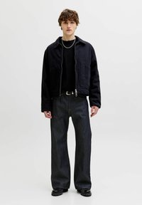 Ung mand med krøllet hår iført en sort lynlåsjakke, sort skjorte, vide mørke jeans med bælte, sorte støvler og et sølvfarvet kædehalskæde mod en hvid baggrund.