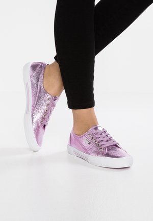 Person iført skinnende metalliske pink sneakers med hvide såler, stående på ét ben, iført sorte bukser mod en hvid baggrund.