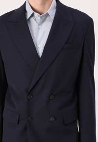 Marineblauwe dubbelknopen blazer met een gelapte kraag, twee voorzakken en zwarte knopen, gedragen over een lichtblauw overhemd met knopen.