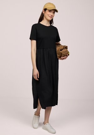 Ung kvinde i sort midi-kjole, beige kasket, hvide sneakers, holder en brun clutch-taske, står mod en ensfarvet baggrund.