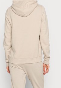 Osoba v béžové mikině a odpovídajících teplácích, viděná ze zadu, stojící na pozadí v jednobarevném světlem.