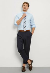 Camicia celeste con bottoni, pantaloni eleganti blu navy, cintura e scarpe marroni, cravatta a righe. Tessuto liscio, taglio sartoriale, leggero risvolto sulle maniche.
