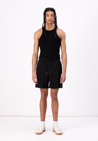 Canottiera nera senza maniche, pantaloni corti neri e sneakers bianche. Il modello sta in piedi, mostrando un look semplice, atletico e casual.