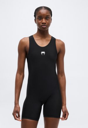 Γυναίκα που φοράει μαύρο αμάνικο αθλητικό κορμάκι με λευκό λογότυπο κόμπρας στο στήθος, που στέκεται μπροστά σε απλό ανοιχτόχρωμο φόντο.