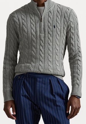 Homme portant un pull gris en maille torsadée avec zip quart, associé à un pantalon bleu marine à fines rayures, mains détendues sur les côtés.