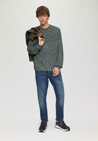 Lichtgrijze gebreide trui met een textuurafwerking, gedragen met blauwe jeans en zwarte sport schoenen; houdt een olijfgroene jas over de schouder.