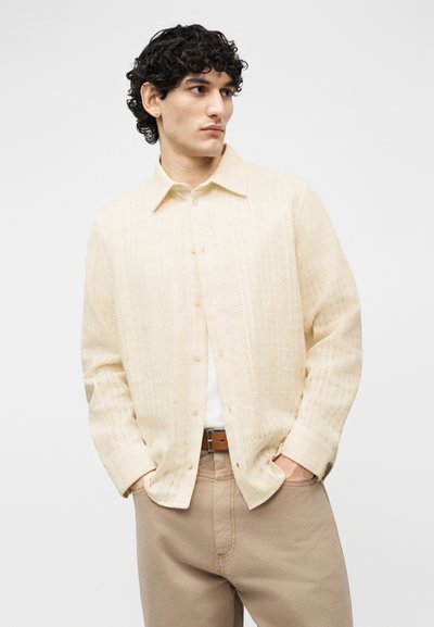 Νεαρός άνδρας με μαύρα σγουρά μαλλιά, φοράει μπεζ υφασμάτινο πουκάμισο με κουμπιά, λευκό εσωτερικό πουκάμισο και ανοιχτό καφέ παντελόνι, με τα χέρια στις τσέπες, κοιτάζοντας δεξιά.