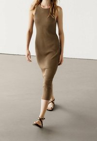 Robe midi marron côtelée et sans manches, ajustée au corps, arrivant mi-mollet. Associée à des sandales plates à lanières dans une couleur complémentaire.