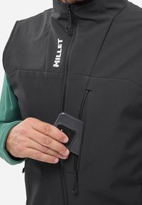 Schwarweste mit Reißverschluss und isoliert, ausgestattet mit einem hohen Kragen und einem Logo. Die Person steckt ein Telefon in eine Tasche.