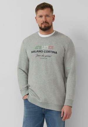 Mann mit Bart, trägt graues Sweatshirt mit dem Text „ATHLETICS MILANO CORTINA Join the games!“ und blaue Jeans, steht vor hellgrauem Hintergrund.