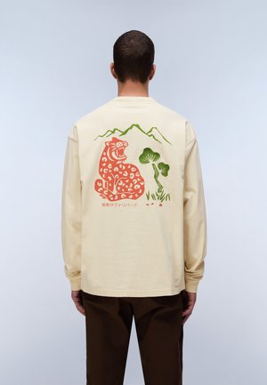 Hombre con camisa beige de manga larga con diseño de leopardo rojo, árboles verdes y montaña en la espalda, combinado con pantalones marrón oscuro.