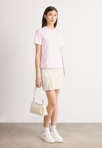 Lyserød bomulds t-shirt, løs pasform, med hvidt logo; lys beige nederdel, med foldedetalje; hvide sneakers; lille cremefarvet håndtaske med rem.