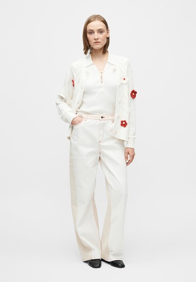Valkoinen neulottu neuletakki, jossa punaisia ja vaaleanpunaisia kukka-aiheisia yksityiskohtia, puettu valkoisen resoripaidan päälle, yhdistetty leveälahkeisiin kermaan sävyisiin housuihin ja mustiin kenkiin.