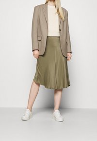 Beige blazer over een witte gebreide top, gecombineerd met een olijfgroene satijnen rok en witte sneakers. De outfit heeft een ontspannen silhouet.