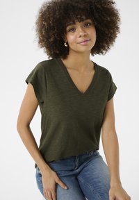 V-apsaugos, trumpomis rankovėmis marškiniai tamsiai alyvuogių spalvos audinyje. Minkštas tekstūros paviršius su subtiliu melange raštu. Derinama su mėlynomis džinsinėmis kelnėmis.