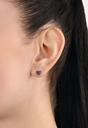 Orecchini a forma di cuore realizzati in gemme viola, montati su una base metallica, esposti su un lobo dell'orecchio con capelli lisci e scuri.