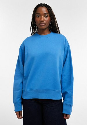 Kvinde med langt fletninger iført en ensfarvet blå, oversize sweatshirt og mørke bukser mod en hvid baggrund.