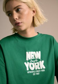 Zelena dukserica s okruglim izrezom; sadrži bijeli natpis "NEW YORK Bagels Autentična Ulična Hrana EST 1997." Mekana tkanina.