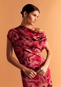 Czerwona i różowa sukienka tie-dye z drapowanym fasonem na ramionach, miękka tekstura, dopasowany krój, uzupełniona o złote kolczyki łańcuszkowe na neutralnym tle.