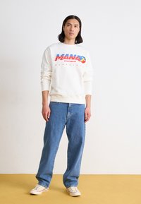 Sweatshirt blanc avec le texte de logo rouge et bleu "MANO SPORTSWEAR", associé à un jean bleu ample et des baskets blanches.