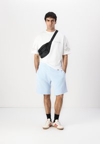 Bílý tričko s dekorativním prošitím, světle modré kraťasy, černá crossbody taška a bílé tenisky s červenými pruhy. Hladké textury látky.