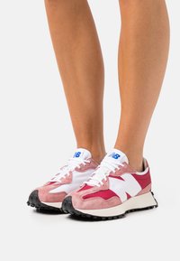 Chaussures de sport avec une tige en daim rose et blanc, une semelle en caoutchouc noire et un logo bleu. Profil bas avec un design à lacets et des accents texturés.