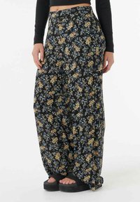 Pantalons larges à fleurs en tissu noir, ornés de petites fleurs bleues et jaunes. Taille élastique ; portés avec des sandales noires.