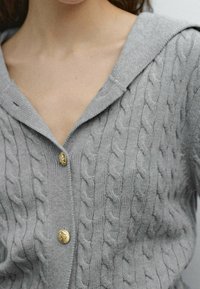 Gray cable-knit cardigan with a hood, featuring textured patterns and gold-tone buttons at the chest. Soft material with a relaxed fit.