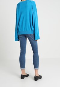 Helles blaues, übergroßes Sweatshirt mit langen Ärmeln, kombiniert mit eng anliegenden Jeans und schwarzen flachen Schuhen, von hinten betrachtet.