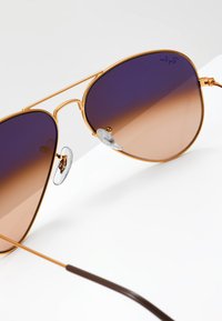 Ochelari de soare aviator aurii cu lentile gradient de violet la chihlimbar. Au tampoane nazale transparente și un braț subțire de culoare maro.